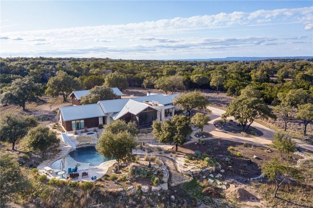 Hill Country ranch house with a metal roof and curved pool, live oaks and a caliche drive under a wide wooded horizon