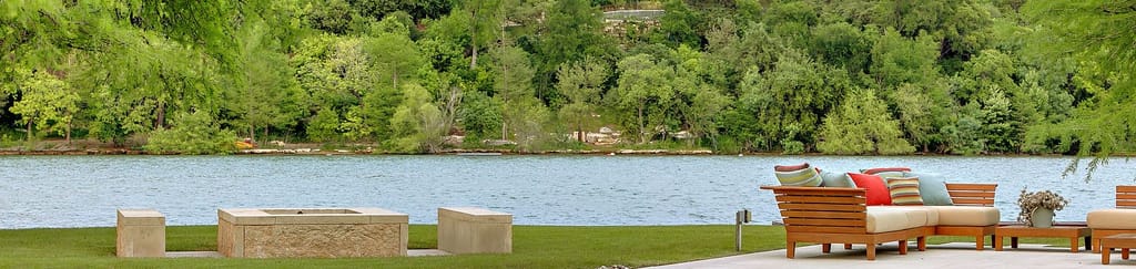 A limestone fire pit on a mown lawn above Lake Austin, teak seating at the pool edge and a wooded far bank